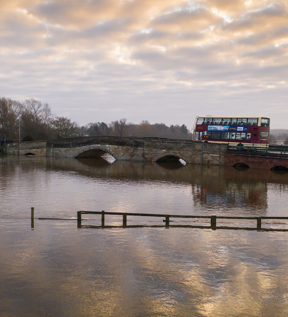 Bus crossing a bridge over a river that has burst its banks and is surrounded by floodwater