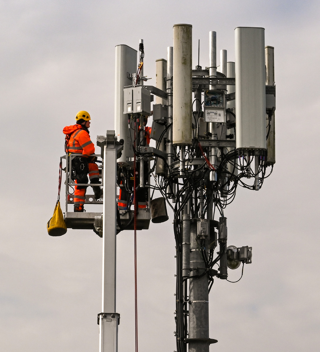 Two male engineers in hi-vis clothing on a cherry picker carrying out maintenance work at the top of a telecoms tower which has approx six rectangular shaped antennas on it
